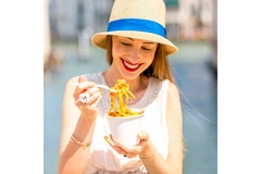 Smiling woman in a straw hat eating noodles from a takeout cup outdoors on a sunny day.